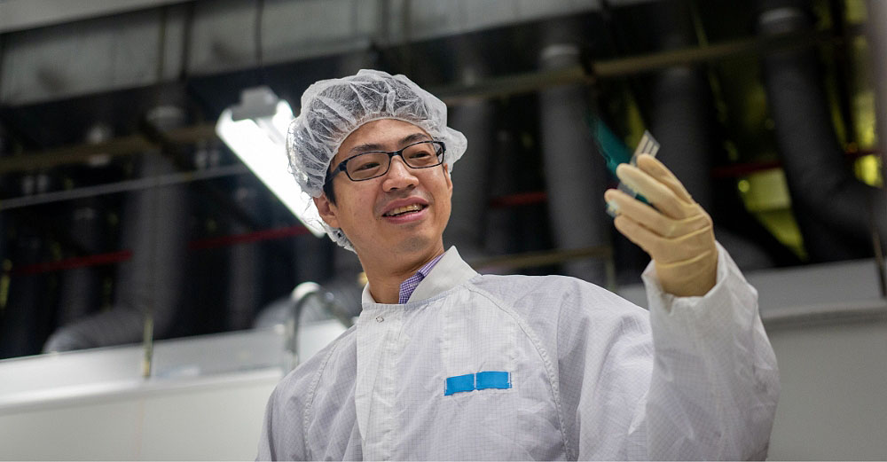 Man in a white labcoat, hair net, and gloves studies object in his hand and smiling