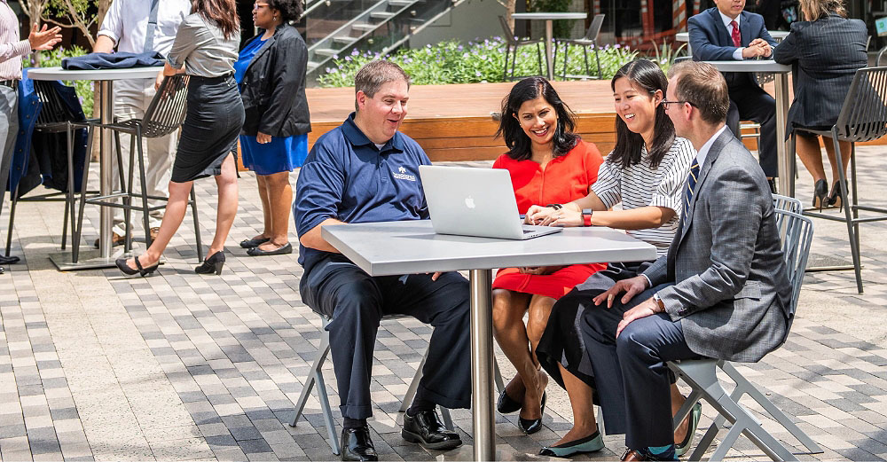 A diverse group of people sit outside at a table looking at a computer and smiling