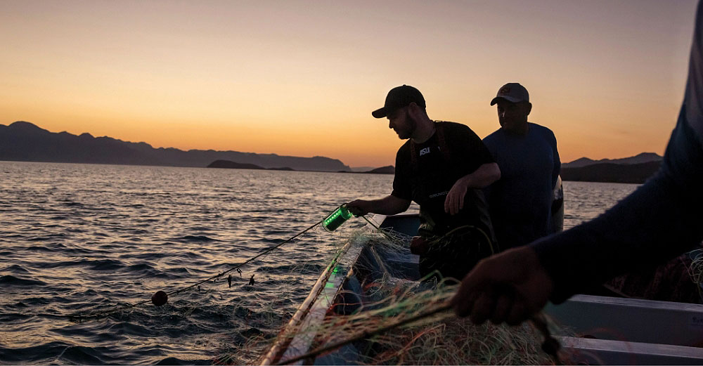 Two men sit on a boat with nets and fishing wire in a big body of water