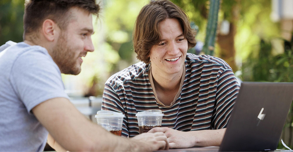 Two male students smiling and looking at laptop computer while sitting outside