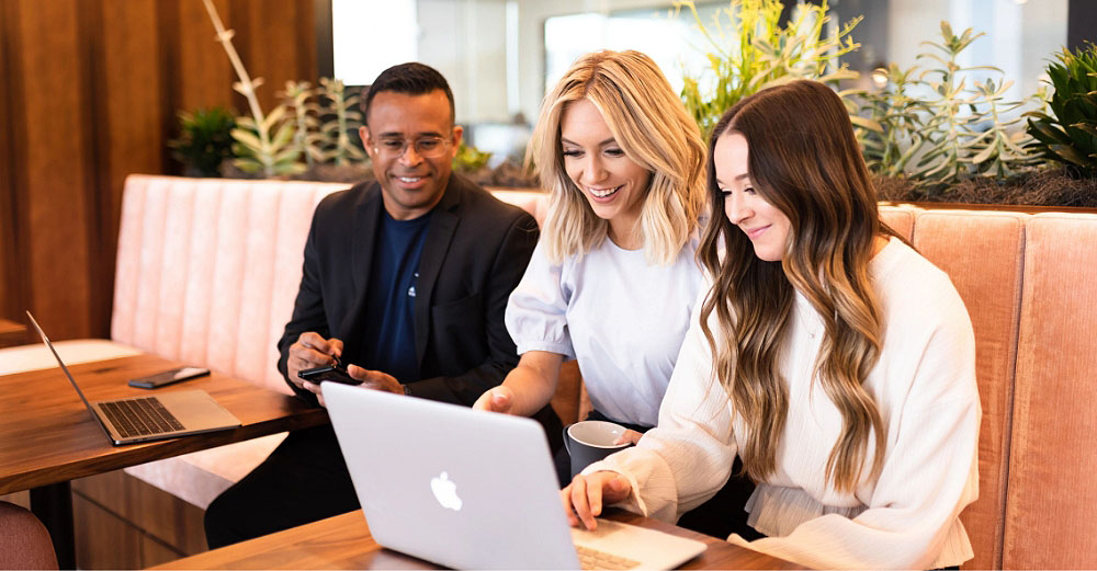 Three people sitting on a bench smiling and looking at a laptop computer