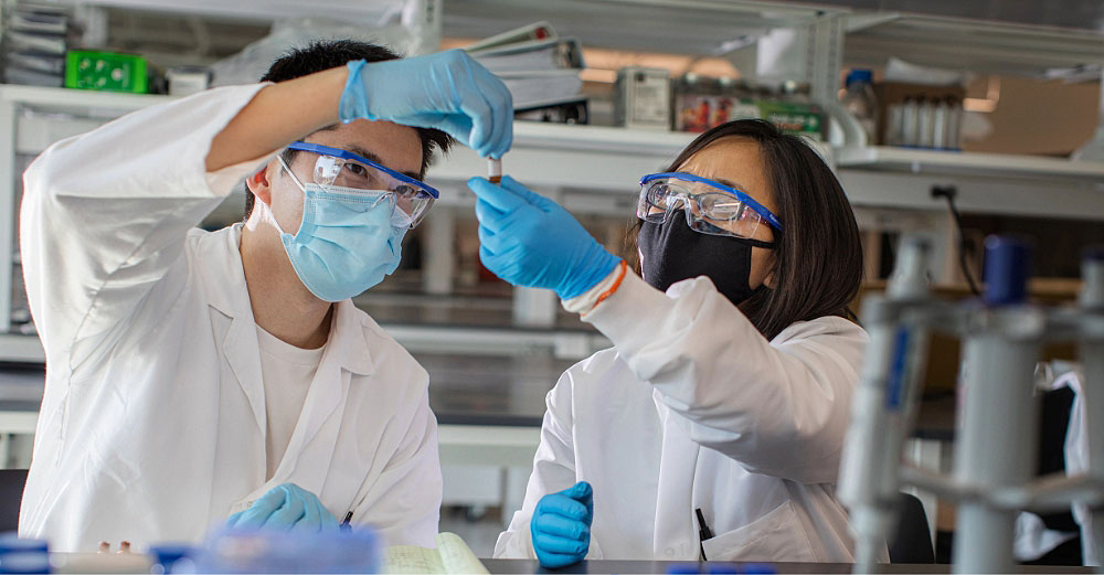 Two students wearing lab coats, masks, goggles, and gloves are exchanging a vial