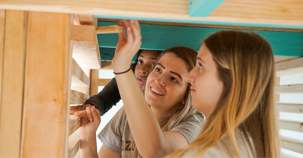 Two similing female students building a wooden structure outside
