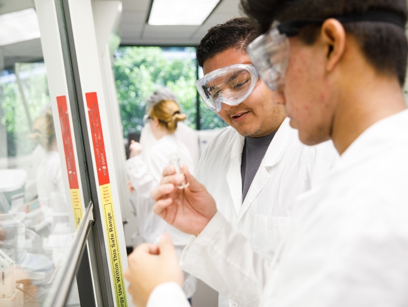 Two male students with lab coats and goggles study a vial